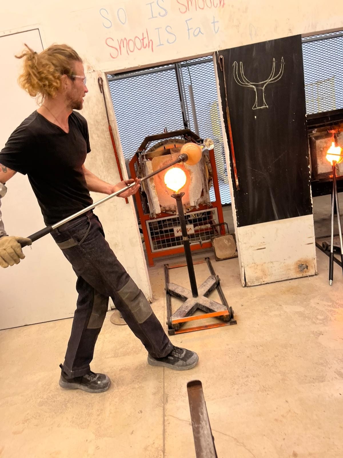 Alex in the studio, carrying a glowing gather of glass on the blowpipe